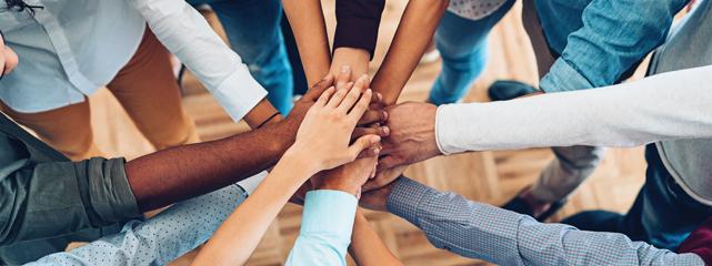 eight people from differnt racial and ethnic groups' arms pointed to a central point with their hands are stacked upon eachother as a team.